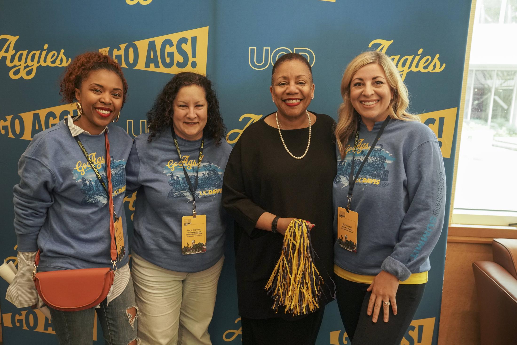 Four women smile at the camera in front of a "Go Ags" backdrop.