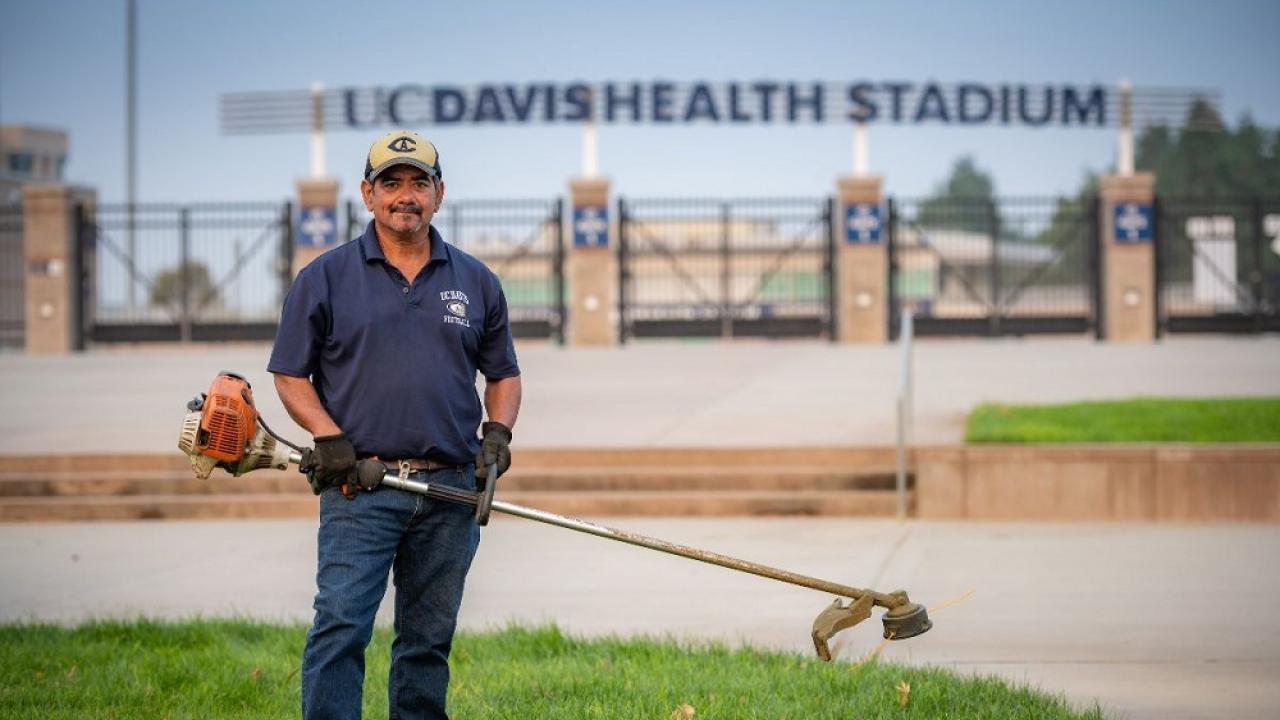 UC Davis Grounds employee in front of UC Davis Health Stadium with a weedwhacker in hand. 