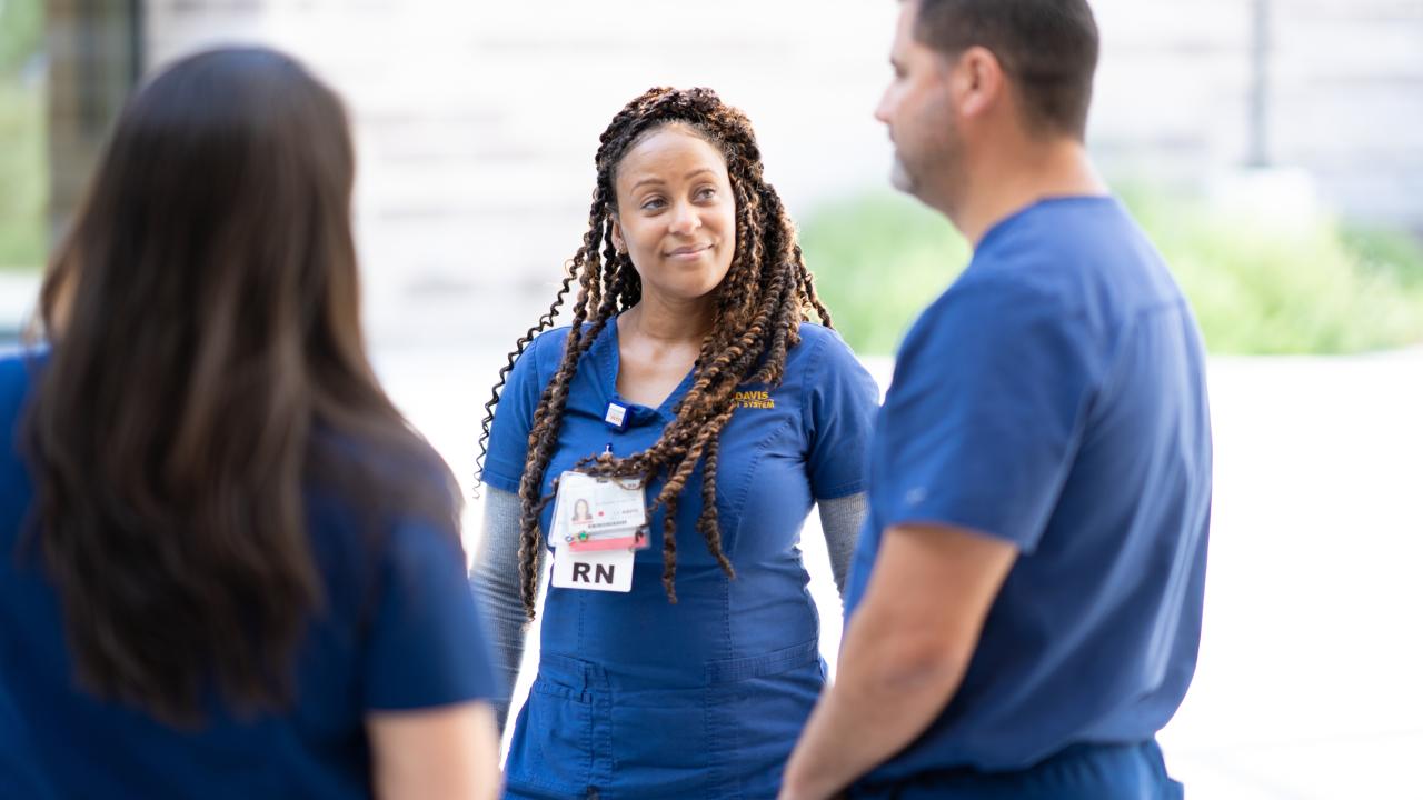 Three nurses stand together. Two have their back facing the camera, while the third looks to one of the other nurses.