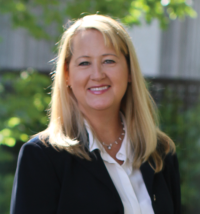 Blonde woman in suit stands in front of greenery