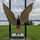 Person standing in front of large golden wings sculpture, with lake and cloudy sky in background.
