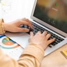 Close-up of hands typing on laptop at desk with colorful chart and glass of water.